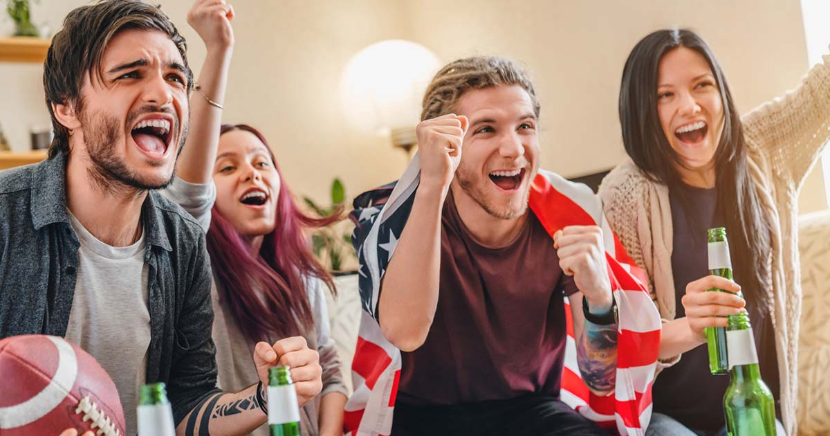 4 millenials cheering while watching a football game. one of them is wrapped in an american flag and another holds a football. there are beers in front of them in green bottles